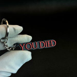 Puede incluir: Un llavero negro con la inscripción "YOU DIED" en rojo. El llavero está hecho de plástico y tiene un anillo de metal plateado.