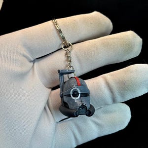 Pode incluir: Um pequeno chaveiro detalhado com um design de capacete cinza com detalhes em preto e uma faixa vermelha. O chaveiro está preso a um anel prateado. Apresentado em uma luva branca.