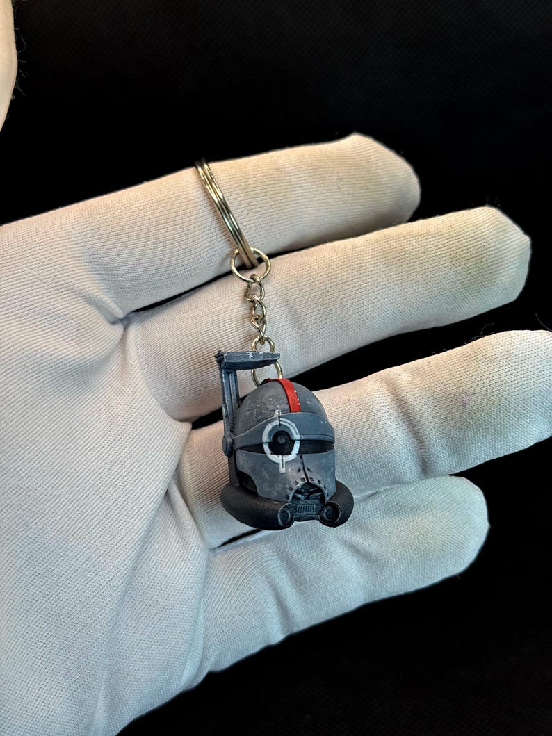 May include: A small, detailed keychain featuring a helmet design with a red stripe. The helmet is primarily gray with black accents and a silver key ring. The keychain is held against a white glove.