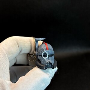 May include: A miniature helmet keychain in shades of gray, black, and red. The helmet features a distinctive design with a red stripe and intricate details. The keychain is attached to a silver ring.