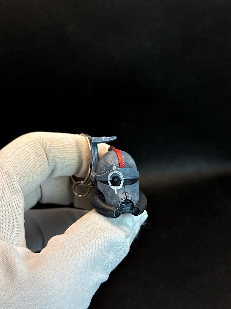 May include: A miniature helmet keychain in shades of gray, black, and red. The helmet features a distinctive design with a red stripe and intricate details. The keychain is attached to a silver ring.