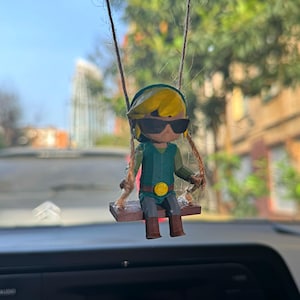 Puede incluir: Una figurita verde y amarilla de un personaje con gafas de sol y un sombrero verde, sentado en un columpio de madera marrón. La figurita está colgada del espejo retrovisor de un coche.