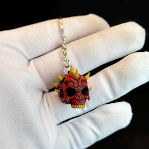 May include: A red and black skull pendant with yellow horns and teeth, attached to a silver chain. The skull has intricate details and is held against a white background.