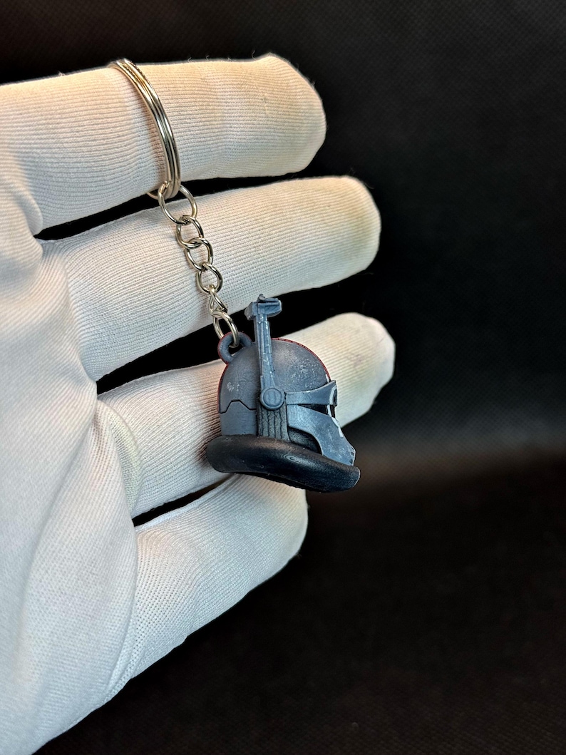 May include: A blue and gray keychain featuring a helmet design. The helmet has a visor and antenna-like details. The keychain is attached to a silver metal ring and chain. The background is dark.