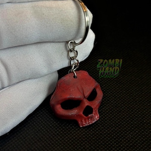 Op de afbeelding: Een rode schedel sleutelhanger met een zilveren sleutelring en ketting. De schedel heeft grote oogkassen en een dreigende uitdrukking. De tekst "ZOMBI HAND" is zichtbaar in het groen.