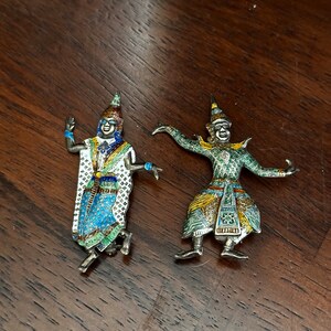 Puede incluir: Dos figuras ornamentadas en tono plateado de bailarines tradicionales. Las figuras están detalladas con esmalte colorido, con detalles en azul, verde, blanco y dorado. Una figura está en pose de salto, la otra con los brazos extendidos. Sobre una superficie de madera.