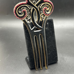 Puede incluir: Un peine para el cabello negro con una parte superior decorativa con un diseño de filigrana dorada y pedrería roja. El peine tiene tres púas y se exhibe sobre un soporte negro sobre un fondo oscuro.
