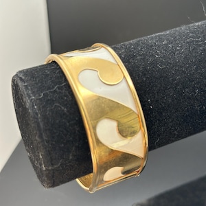 May include: Gold-tone cuff bracelet with a white enamel inlay featuring a repeating wave design. The bracelet has a polished finish and a hinged closure. A stylish accessory for everyday wear.