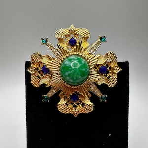 May include: A gold-toned brooch with a green stone center surrounded by blue and clear rhinestones. The brooch is in the shape of a starburst with intricate details.