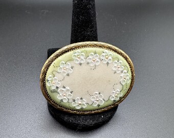 May include: Oval-shaped brooch with a green background and white flowers. The brooch is surrounded by a gold-coloured metal frame.