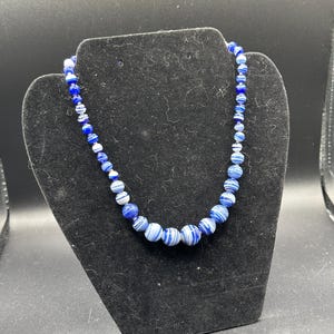 Puede incluir: Un collar de cuentas con esferas en varios tonos de azul y blanco. Las cuentas presentan un patrón jaspeado y arremolinado, con las cuentas más grandes situadas en el centro y las más pequeñas disminuyendo hacia los extremos.