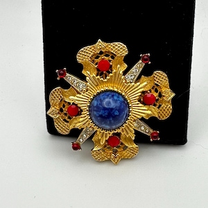 May include: A gold-toned brooch with a blue stone in the center and red and clear rhinestones around it. The brooch is in the shape of a starburst.