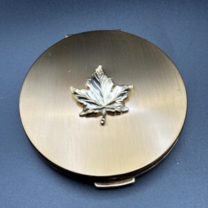 Peut inclure: Un poudrier rond de couleur dorée avec un emblème de feuille d'érable argenté en relief. Le poudrier a un design simple et élégant et une fermeture à fermoir. La surface a une finition brossée, et l'esthétique générale est vintage.