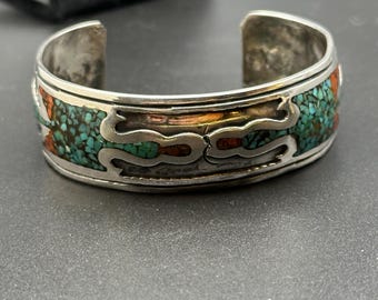 May include: A silver bracelet with turquoise and coral inlay. The bracelet has a geometric design and is made by a Native American artist.