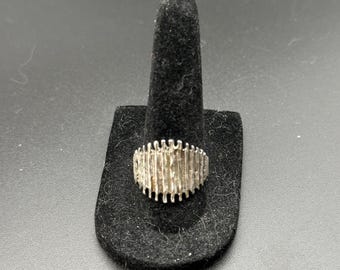 May include: A silver ring with a textured, rectangular design. The ring is displayed on a black ring holder.