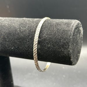 May include: A thin, circular silver-toned bangle bracelet featuring a continuous, repeating diagonal etched pattern along its entire outer surface. The metal has a polished, reflective finish that catches the light, highlighting the intricate texture.