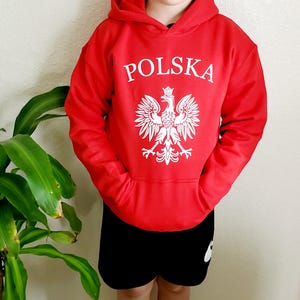 May include: A red hooded sweatshirt with the word "POLSKA" in white block letters above a white Polish eagle emblem. The sweatshirt has a front pocket and is paired with black shorts.