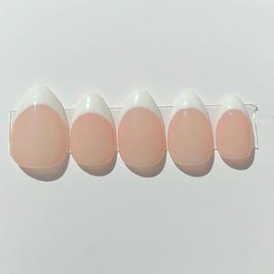 May include: Five oval-shaped, artificial nails with a classic French manicure design. Each nail features a pale pink base with a white tip. The nails are arranged in a row on a clear adhesive strip.