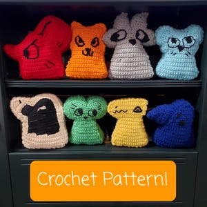 Op de afbeelding: Een plank met acht gehaakte knuffels. De knuffels zijn een rood monster, een oranje kat, een grijze wasbeer, een blauwe kat, een zwart-wit monster, een groen monster, een geel monster en een blauw monster. De plank is gelabeld met "Crochet Pattern!"