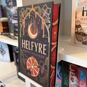 May include: A hardcover book titled Helfyre by Mariel Pomeroy with a black cover featuring gold foil illustrations of a crescent moon, flames, and citrus slices. The page edges are decorated with a repeating pattern of red and orange citrus slices.