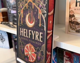 May include: A hardcover book titled Helfyre by Mariel Pomeroy with a black cover featuring gold foil illustrations of a crescent moon, flames, and citrus slices. The page edges are decorated with a repeating pattern of red and orange citrus slices.