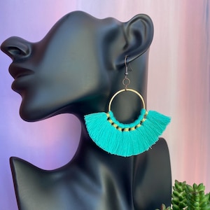 May include: Turquoise tassel earrings with gold-tone hoops. The earrings feature a fan-shaped design with a fringe of turquoise thread and gold accents. The earrings are displayed on a black mannequin head.