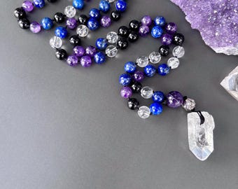 Op de afbeelding: Een kralenketting met een herhalend patroon van ronde, gepolijste amethist, lapis lazuli, zwarte onyx en heldere kwartskralen. De ketting is afgewerkt met een enkele, heldere, zeshoekige kwartskristallen hanger bevestigd aan een zilverkleurige kraal.