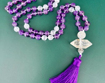 May include: A beaded necklace featuring alternating purple amethyst and clear quartz spheres, accented by a double-terminated clear quartz crystal point, a silver-toned metal bead cap, and a long, vibrant purple silk tassel at the base.