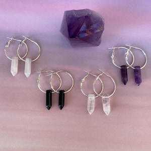 Puede incluir: Una selección de pendientes de aro plateados con colgantes de cristal y piedras preciosas. Los pendientes presentan colgantes de cuarzo transparente, ónix negro y amatista. También hay un gran cristal de amatista púrpura facetado.