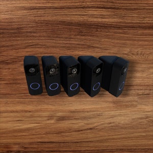 Op de afbeelding: Vijf zwarte, rechthoekige beveiligingscamera's met een lens en een blauwe oplichtende ring aan de onderkant. De camera's staan in een rij op een houten oppervlak. Modern en strak design.