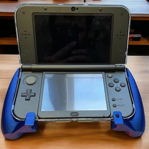 Könnte beinhalten: Handheld-Spielkonsole mit blauem Griffaufsatz. Die Konsole ist geöffnet und zeigt zwei Bildschirme an. Die Tasten und Bedienelemente sind sichtbar, einschließlich des Steuerkreuzes, der Funktionstasten und der Start/Select-Tasten. Die Konsole ist grau und schwarz.