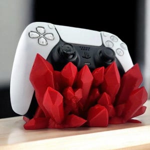 May include: A white and black PlayStation 5 controller rests on a vibrant red crystal-shaped stand. The controller features a sleek design with black joysticks and buttons. The stand adds a decorative touch, enhancing the gaming setup.