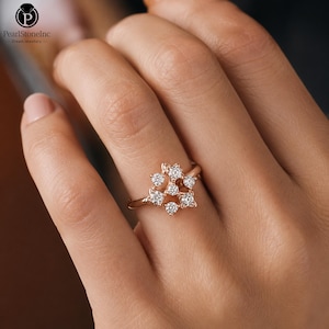 May include: A rose gold ring with a floral arrangement of clear gemstones. The ring is shown on a hand, highlighting the design. The band is slender and the gemstones are round-cut. The ring's design is delicate.