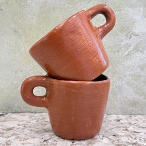 May include: Two stacked, handmade terracotta mugs. The mugs are a warm reddish-brown color with a textured surface and rounded handles. The mugs are empty and ready to be filled with your favorite beverage.