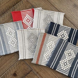 May include: A collection of woven square coasters in various colors, including red, gray, and navy blue. Each coaster features a geometric pattern in white, with some having striped or solid borders. The coasters are arranged on a wooden surface.