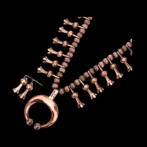 Puede incluir: Un conjunto de joyas en tono cobre que incluye un collar y pendientes. El collar tiene un colgante grande y circular y hebras de cuentas con detalles florales. Los pendientes reflejan el diseño floral. El conjunto se muestra sobre un fondo negro.