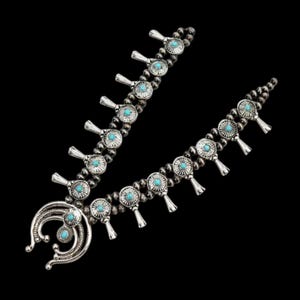 Puede incluir: Un collar de flores de calabaza plateado con detalles de piedras de color turquesa. El collar presenta un colgante en forma de media luna y una serie de flores y cuentas estilizadas. El fondo es negro.