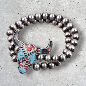 May include: Silver beaded bracelet featuring a decorative bull skull pendant. The pendant has a turquoise, red, black, and white geometric design. The bracelet is made of multiple strands of silver beads.