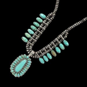 Puede incluir: Un collar de plata con piedras turquesas. El collar presenta un gran colgante ovalado con una piedra turquesa central rodeada de piedras más pequeñas. Tiene una serie de piedras turquesas en forma de lágrima.