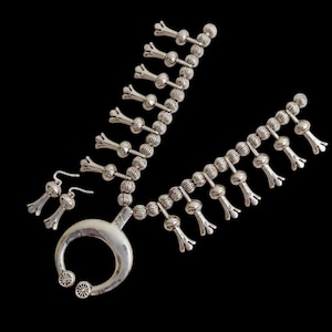 Puede incluir: Un conjunto de joyas plateadas que incluye un collar y pendientes a juego. El collar tiene un colgante grande en forma de media luna y hebras de cuentas con detalles florales. Los pendientes son pequeñas versiones colgantes del diseño floral.