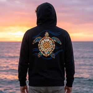 Puede incluir: Una sudadera negra con capucha que presenta en la espalda un gráfico de una tortuga marina compuesta por varias conchas y piedras en tonos beige, marrón y azul. La tortuga está rodeada de patrones de olas azules sobre el tejido negro.