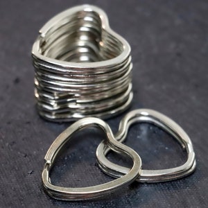 Könnte beinhalten: Ein Stapel silberfarbener herzförmiger Schlüsselanhänger. Zwei Ringe befinden sich im Vordergrund, der Rest ist dahinter gestapelt. Die Schlüsselanhänger sind aus Metall und haben eine glatte, glänzende Oberfläche. Der Hintergrund ist eine dunkle, strukturierte Oberfläche.
