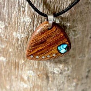 May include: A wooden pendant with a triangular shape, featuring a natural wood grain pattern. The pendant has a small turquoise inlay and a series of small, silver-colored dots along one edge. It hangs from a black cord.