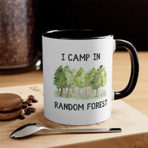 May include: White ceramic mug with a black rim and handle. The mug has a watercolor illustration of a forest with the text "I camp in random forest".