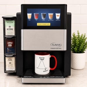 May include: A black and silver Flavia Creation 600 coffee machine with a touchscreen display showing drink options. A white mug with a red interior and handle features a bell curve graphic with the text Don't be mean, Be significant.
