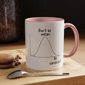 May include: A white ceramic mug with a soft pink interior and handle. The exterior features a black line drawing of a bell curve graph with the text "Don't be mean" above the peak and "Be significant" below the right tail of the curve.