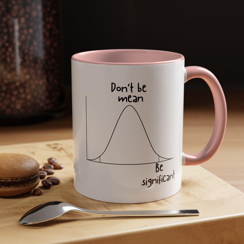 May include: A white ceramic mug with a soft pink interior and handle. The exterior features a black line drawing of a bell curve graph with the text "Don't be mean" above the peak and "Be significant" below the right tail of the curve.