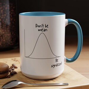 May include: A white ceramic mug with a teal interior and handle. The exterior features a black line drawing of a bell curve graph with the text Don't be mean above the peak and the text Be significant below the right tail of the curve.