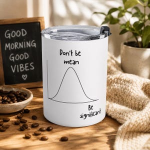 May include: White travel mug with a black lid, featuring the text "Don't be mean, Be significant" with a bell curve graph. The mug is on a wooden surface with coffee beans and a chalkboard that says "Good Morning Good Vibes".