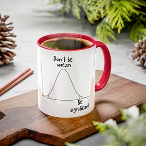May include: A white ceramic mug with a red interior and handle, featuring a black line drawing of a bell curve graph. The text "Don't be mean" is printed above the curve, and "Be significant" is printed below the curve in black lettering.